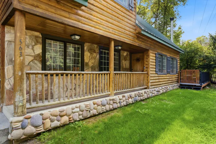 Log cabin exterior featuring a covered porch with natural wood railings, stone accents, and a verdant lawn