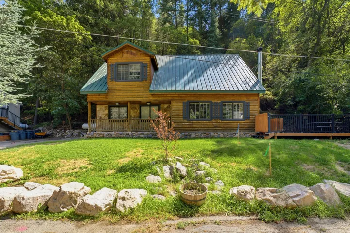 Log cabin exterior with a green metal roof, stone foundation, covered front porch with wood railings, and a side deck with dark railings