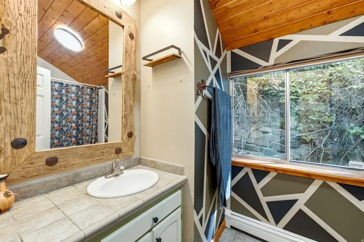Wood-paneled vaulted ceiling, large framed mirror, tile countertop with integrated sink, window with wood sill, and geometric accent wall