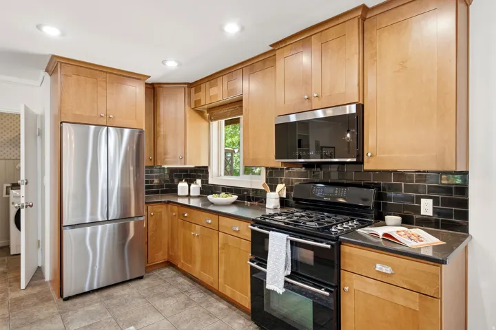 Recessed lighting illuminates wood cabinetry, stainless steel appliances, black tile backsplash, and dark countertops