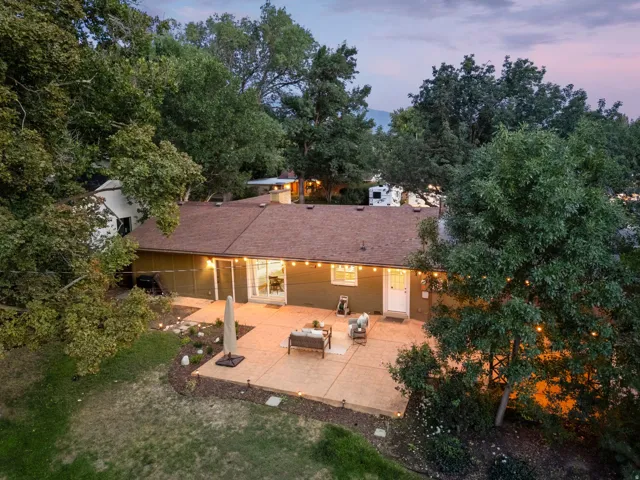 Expansive concrete patio, mature trees, brown shingle roof, green exterior siding, and white trim