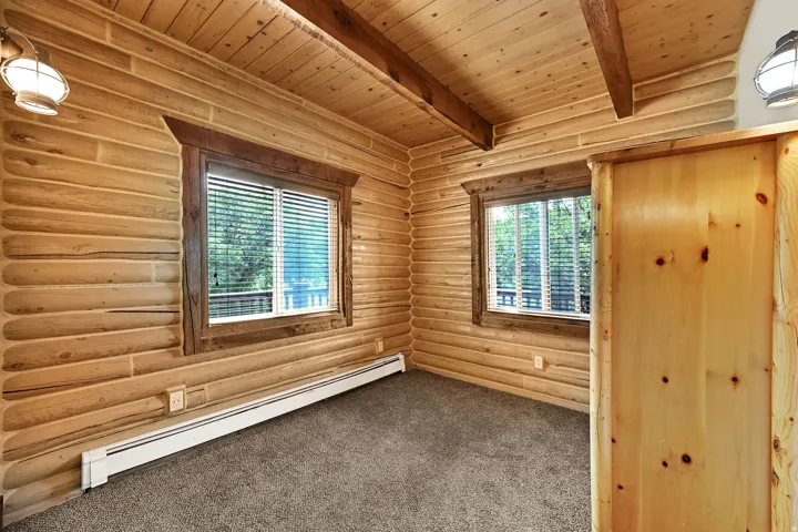 Log wall construction with exposed ceiling beams, two windows with wood trim, and wall-mounted light fixtures