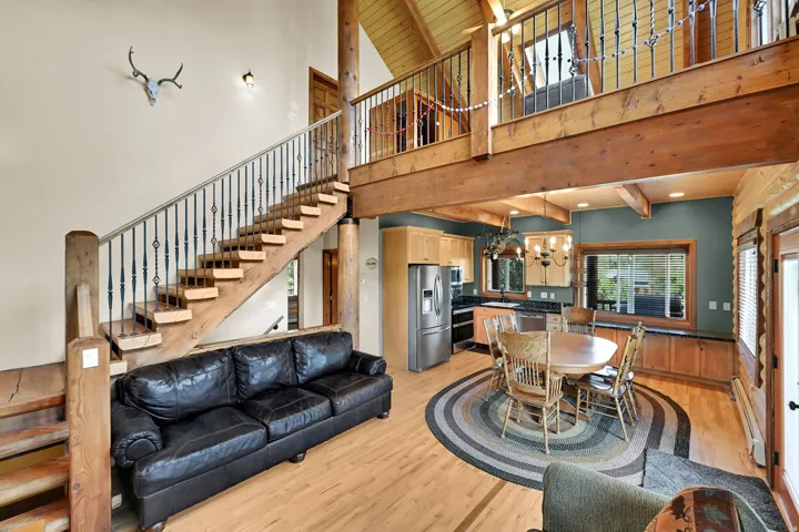 Vaulted ceiling with exposed wood beams, wood-finish flooring, open-concept living area, kitchen with wood cabinetry and stainless steel appliances, and a wood staircase with wrought iron balusters