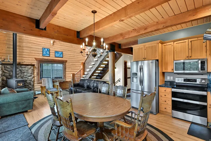 Wood-paneled ceilings with exposed wood beams, stainless steel kitchen appliances, wood cabinetry, stone-clad wood-burning stove, and a wood staircase with wrought iron balusters