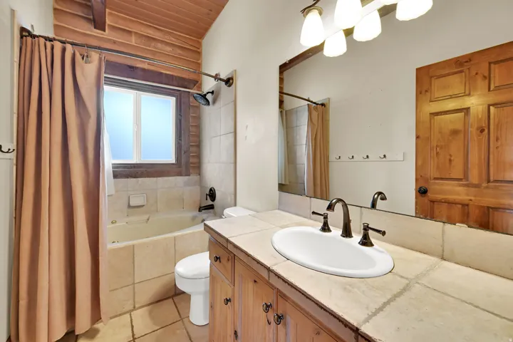 Wood-plank ceiling, tiled bathtub surround with a window, wood-finish vanity with an integrated sink, tiled countertop, and bronze-finish fixtures
