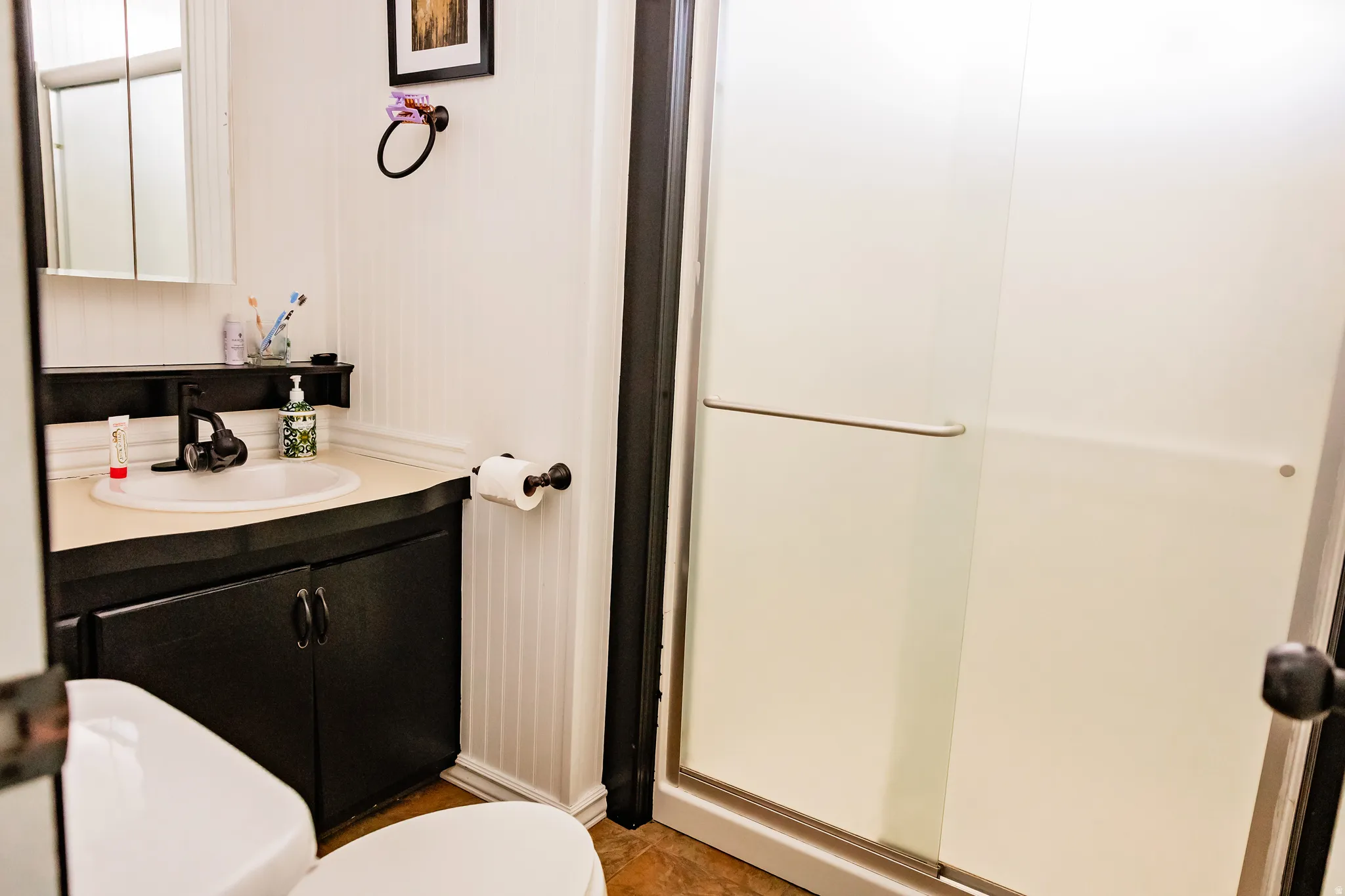Frosted glass shower enclosure, white beadboard wainscoting, black vanity with integrated sink, and wood-finish flooring