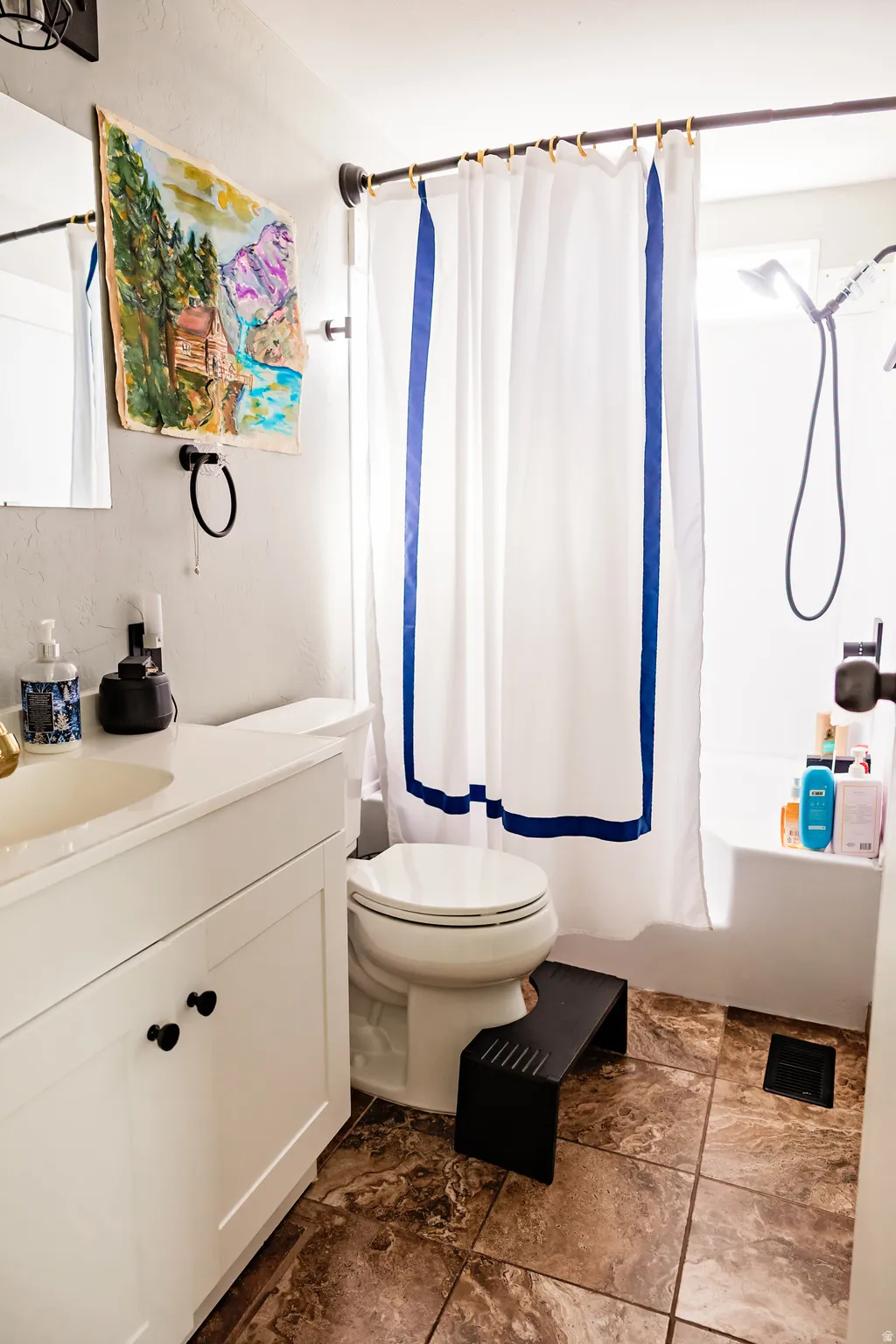 White vanity with black knob hardware, tan and brown patterned floor tiling, and a white shower-over-tub combination