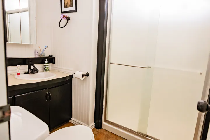 Frosted glass shower enclosure, white beadboard wainscoting, black vanity with integrated sink, and wood-finish flooring