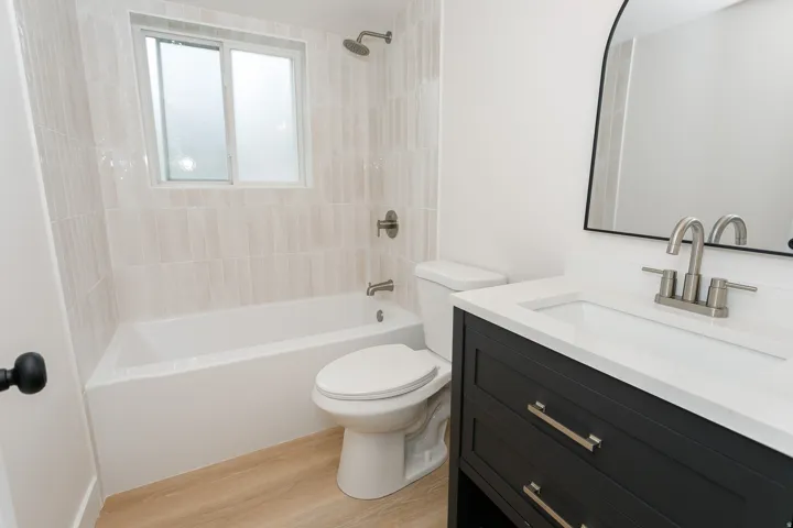 Bathroom featuring a white bathtub, wood-finish flooring, a dark vanity with a white countertop, and a contemporary mirror