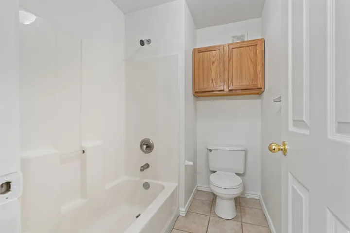 White bath and shower combination with chrome fixtures, tiled flooring, built-in wooden cabinetry, and a white commode