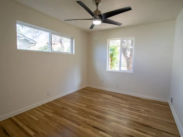 Hardwood flooring with white baseboards, a ceiling fan with integrated lighting, and multiple windows providing natural illumination