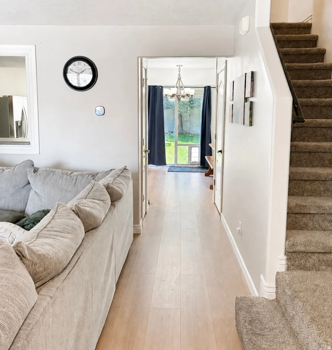 Light wood-finish flooring extending through the hallway, carpeted stairs with a dark handrail, and a contemporary chandelier visible through the doorway