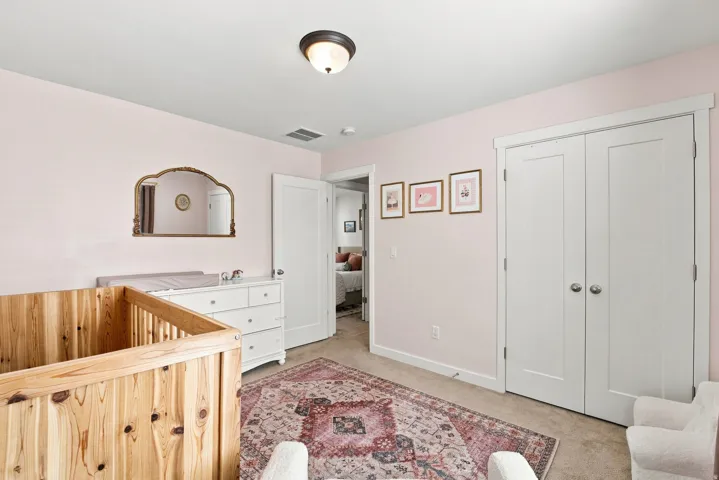 Charming nursery featuring soft pink walls, natural wood crib with spindle detailing, white dresser with integrated changing surface, dual white closet doors, ceiling-mounted fixture, and traditional area rug anchoring the space
