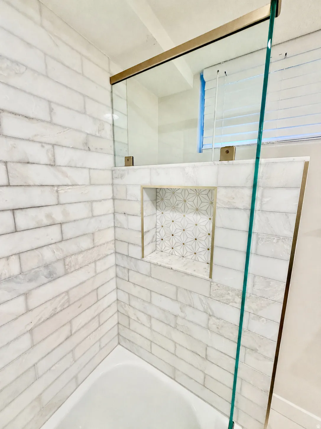 Bathtub with subway tile surround, integrated shower niche with decorative tile, and glass shower enclosure with brass-finish hardware