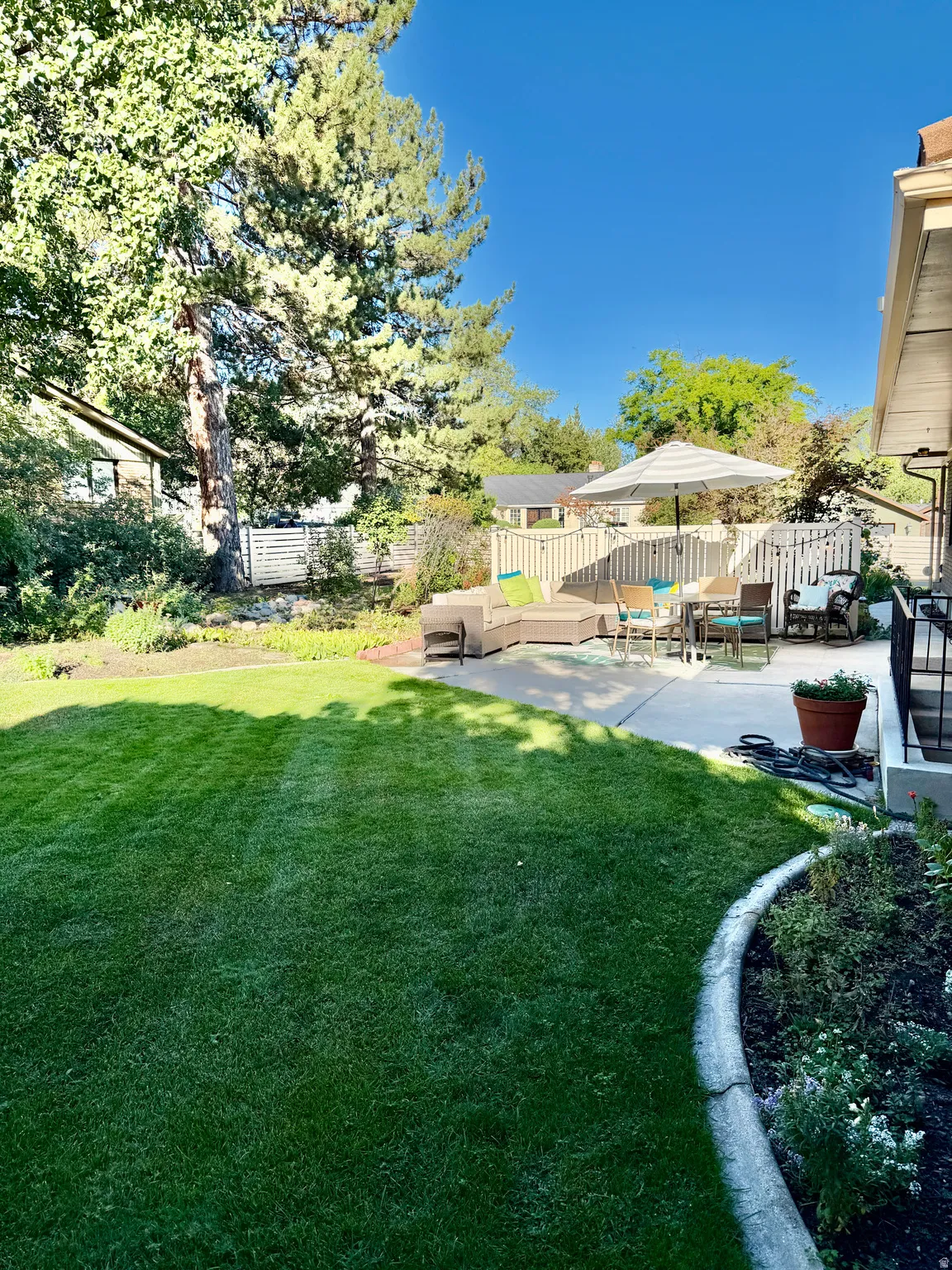 Expansive lawn with a concrete patio, bordered by mature trees and landscaped garden beds
