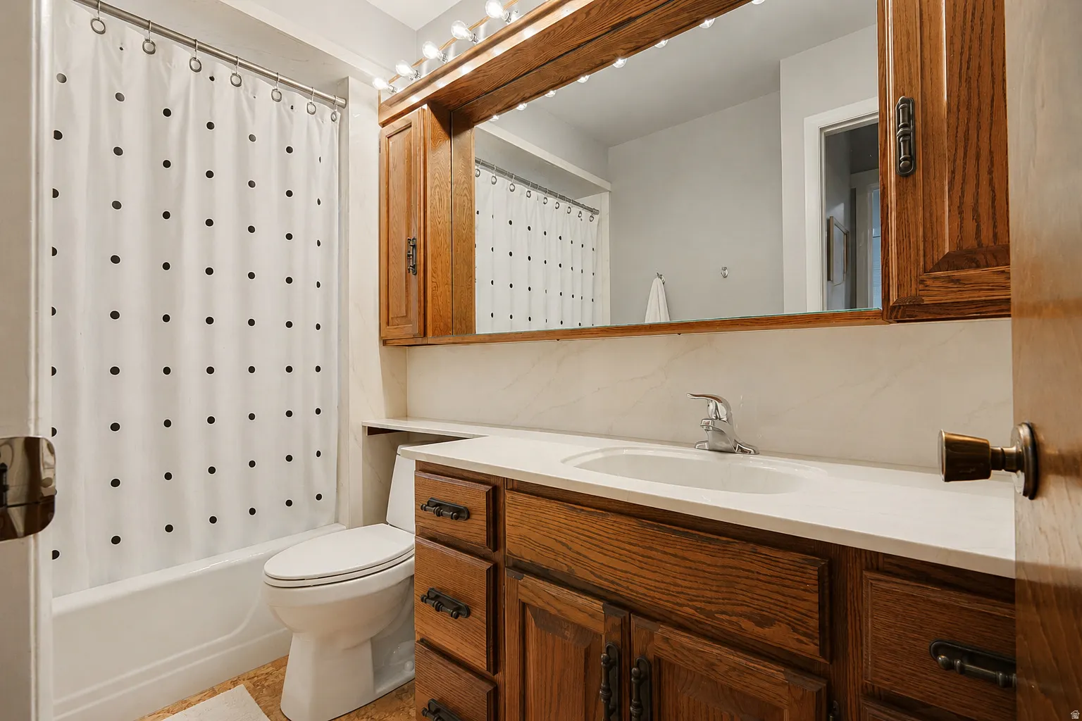 White bathtub with shower, wood-finish vanity with countertop, integrated sink, and wall-mounted mirror with overhead lighting