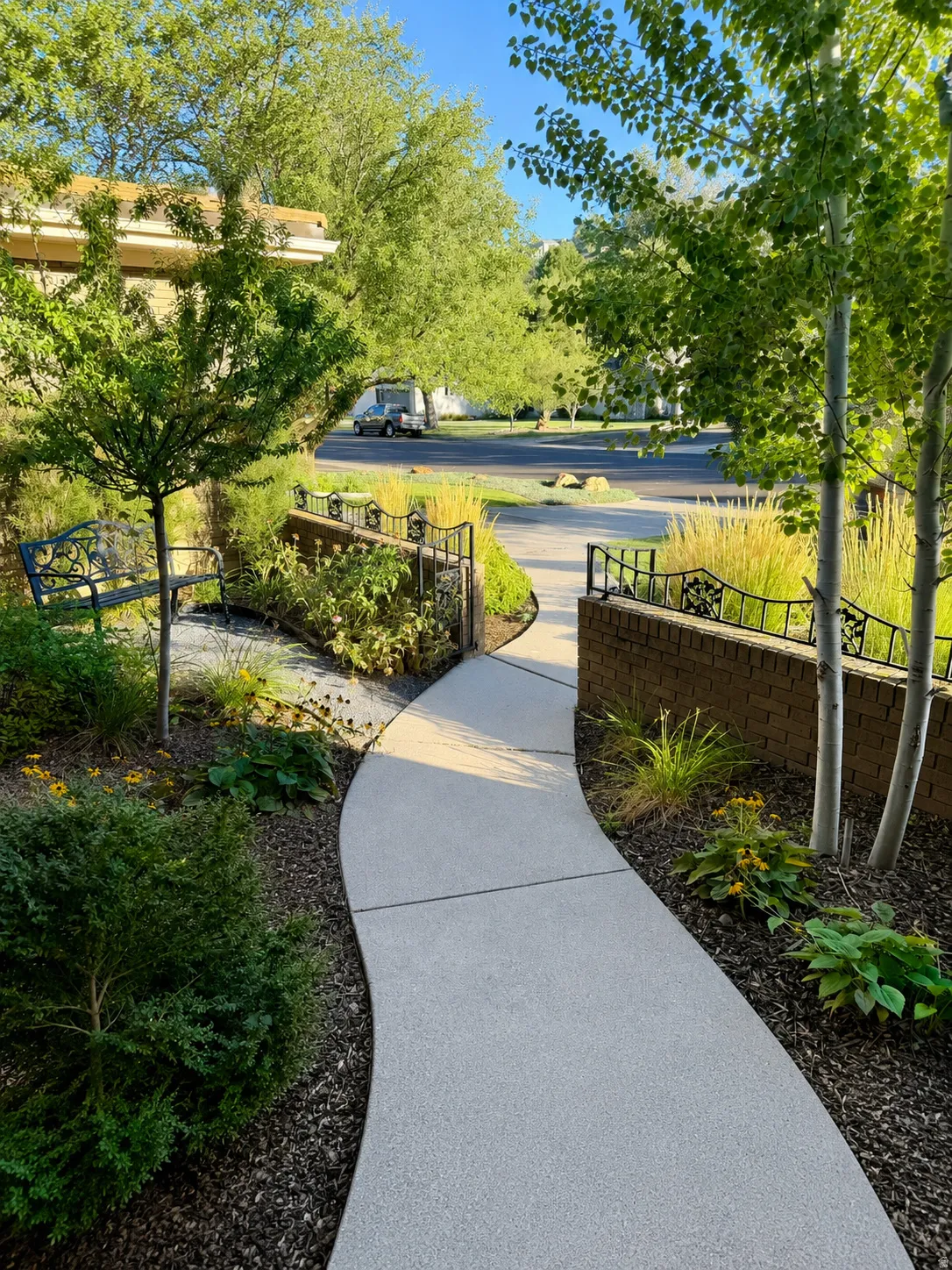 Curved exposed aggregate pathway with manicured landscaping, mature trees, and decorative brick retaining walls with wrought iron accents