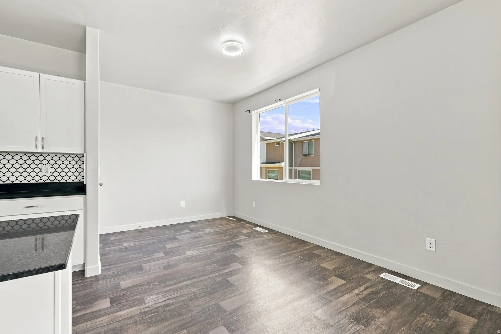 Wood-finish flooring, white cabinetry, dark countertop, patterned tile backsplash, and a window providing natural light