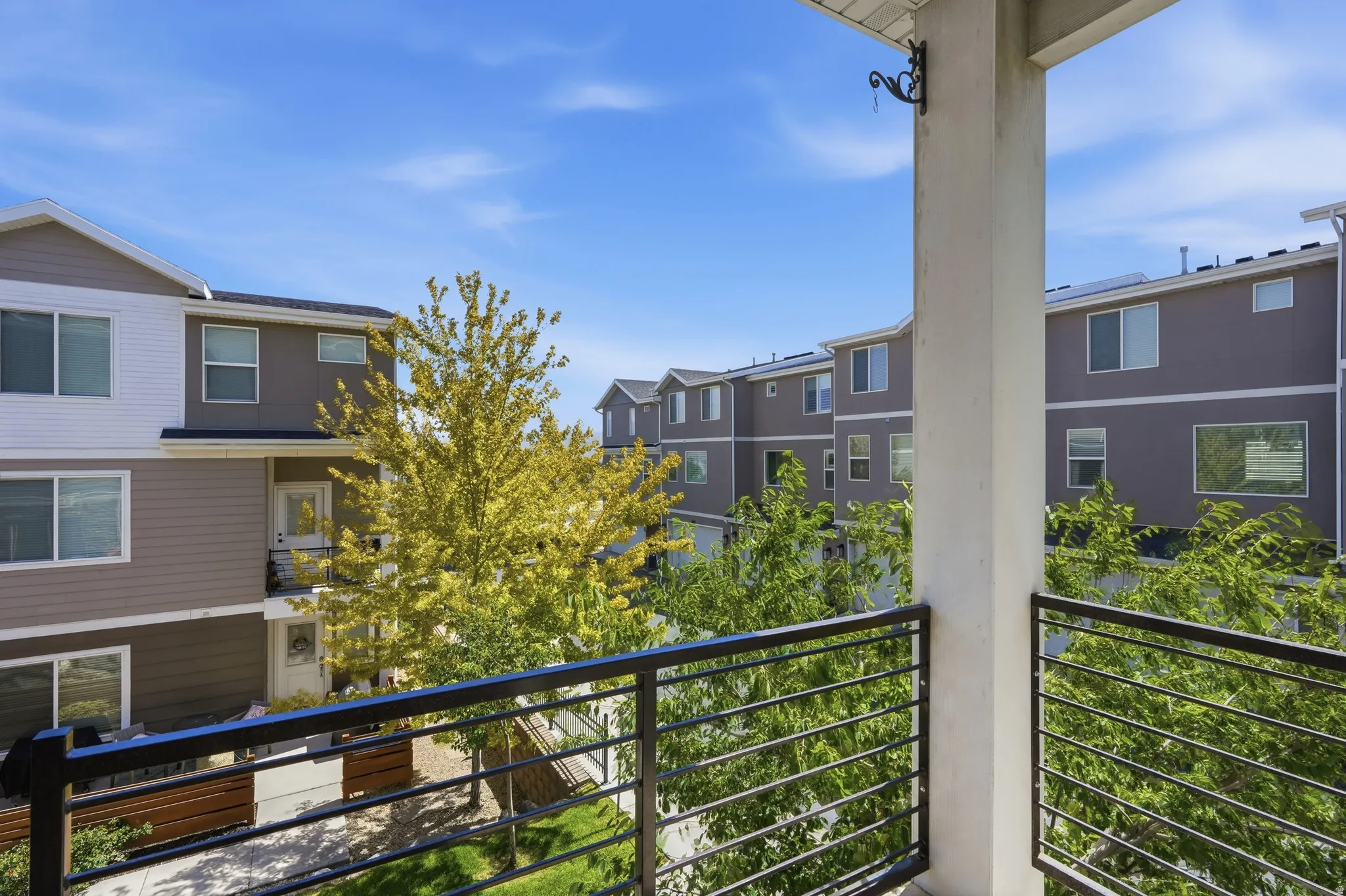 Balcony with black metal horizontal railing overlooking landscaped common areas and multi-story townhomes with light siding