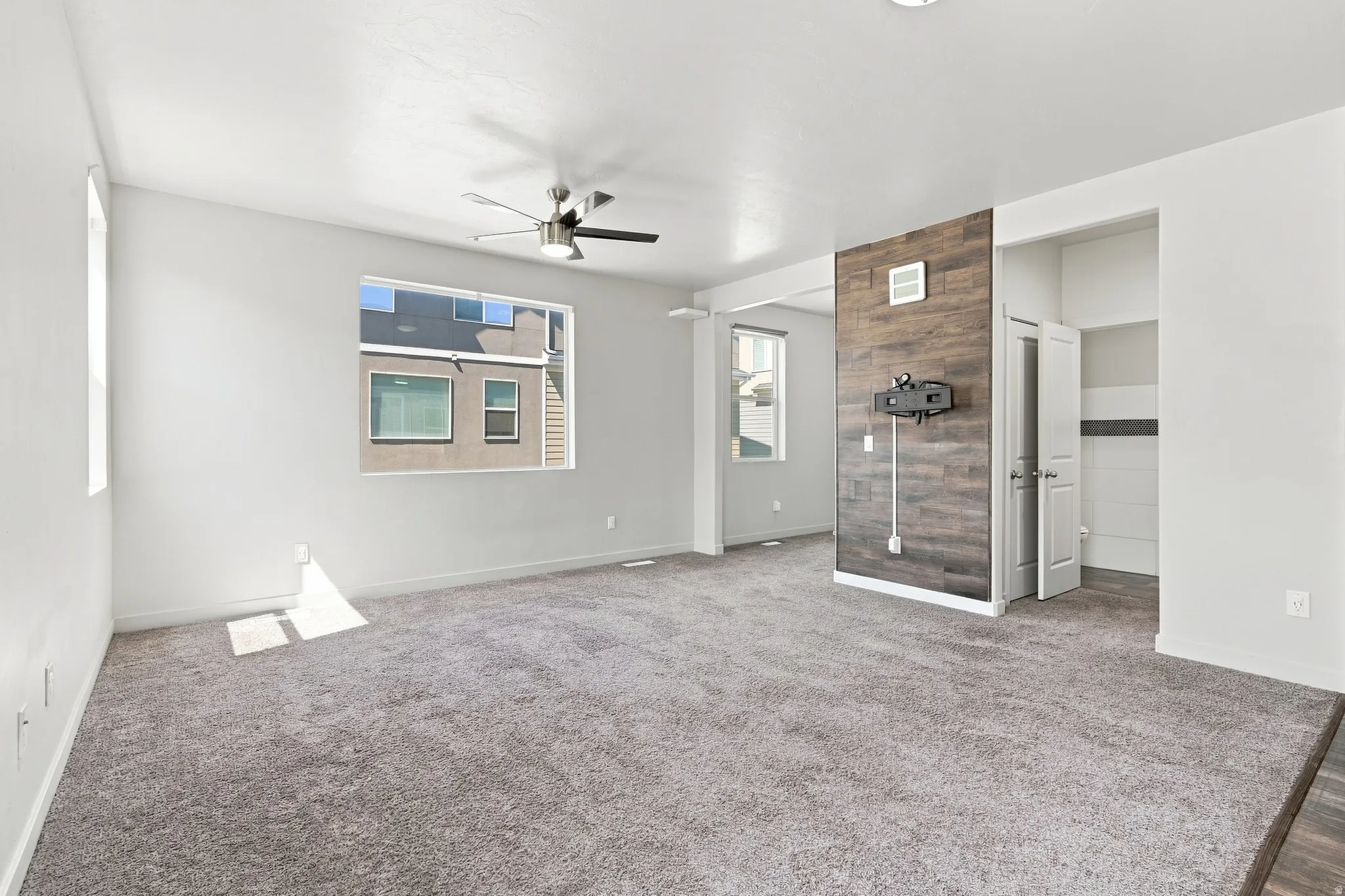Expansive carpeted living space with a ceiling fan, large window, and wood-panel accent wall
