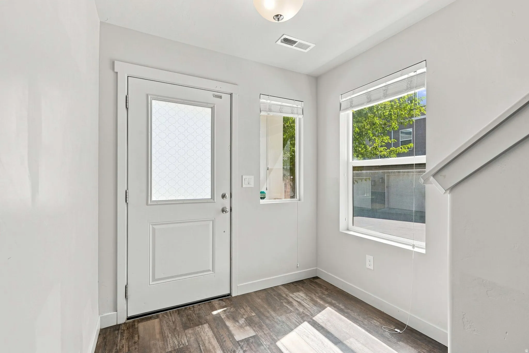 White paneled entry door with decorative glass insert, wood-finish flooring, two windows with blinds, and white walls