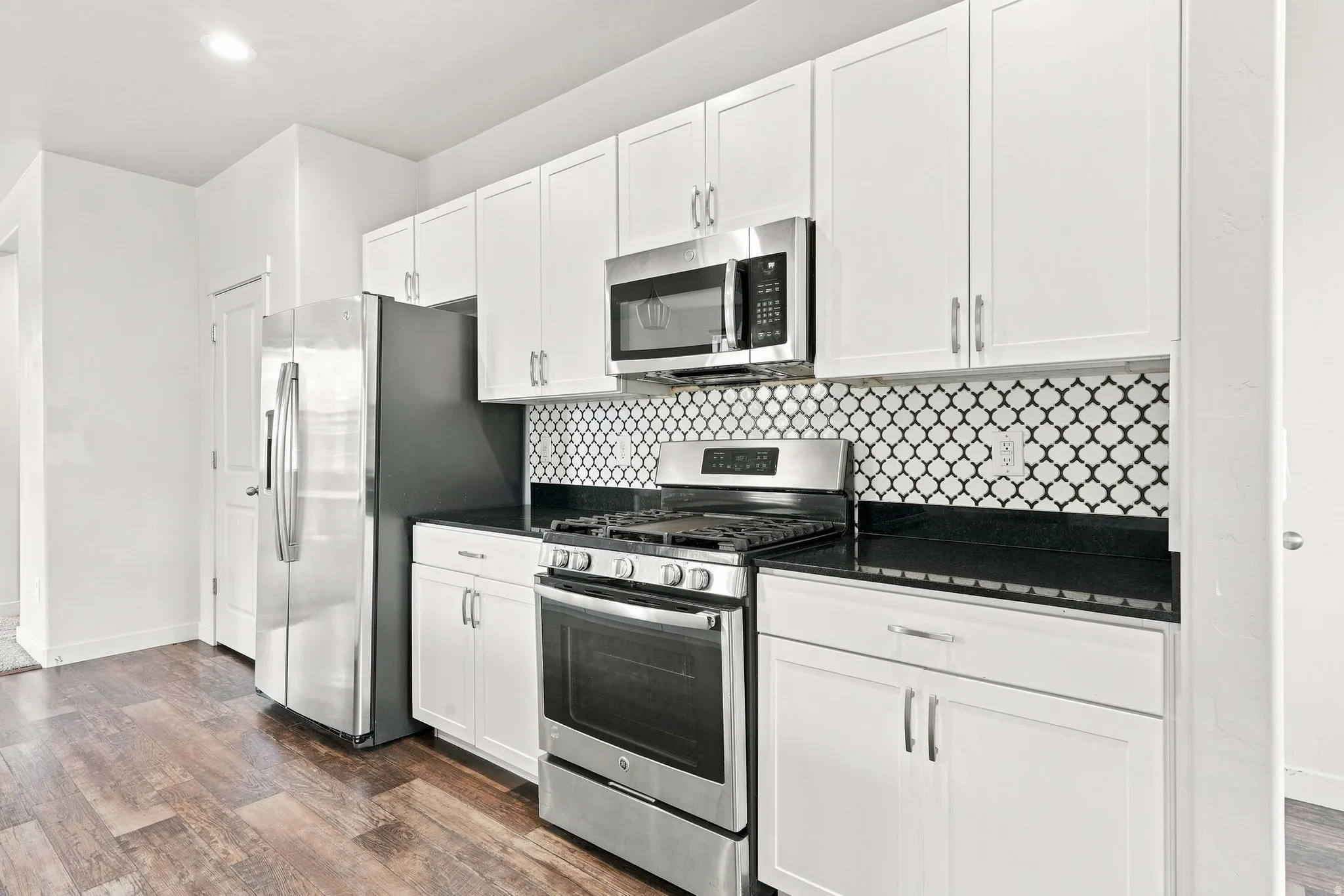 White cabinetry with stainless steel hardware, black countertop, patterned tile backsplash, stainless steel appliances, and wood-finish flooring