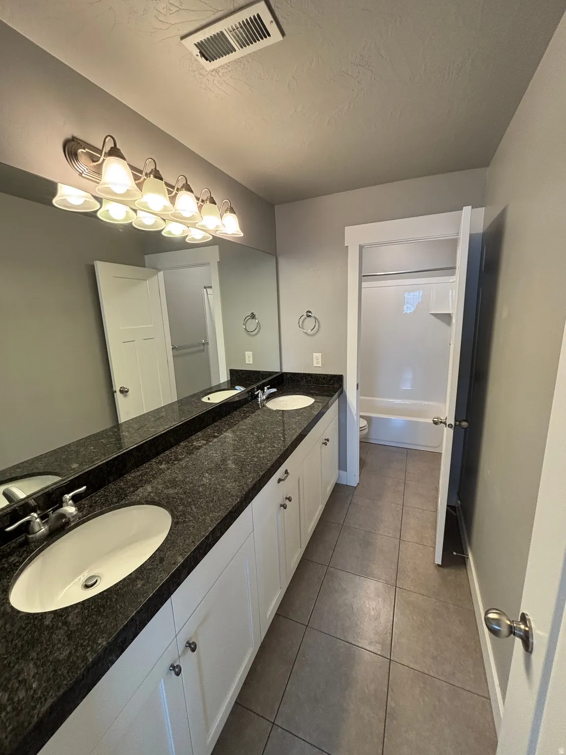 Master bathroom featuring a dual-sink vanity with dark stone countertop, white cabinetry with chrome hardware, and an illuminated mirror
