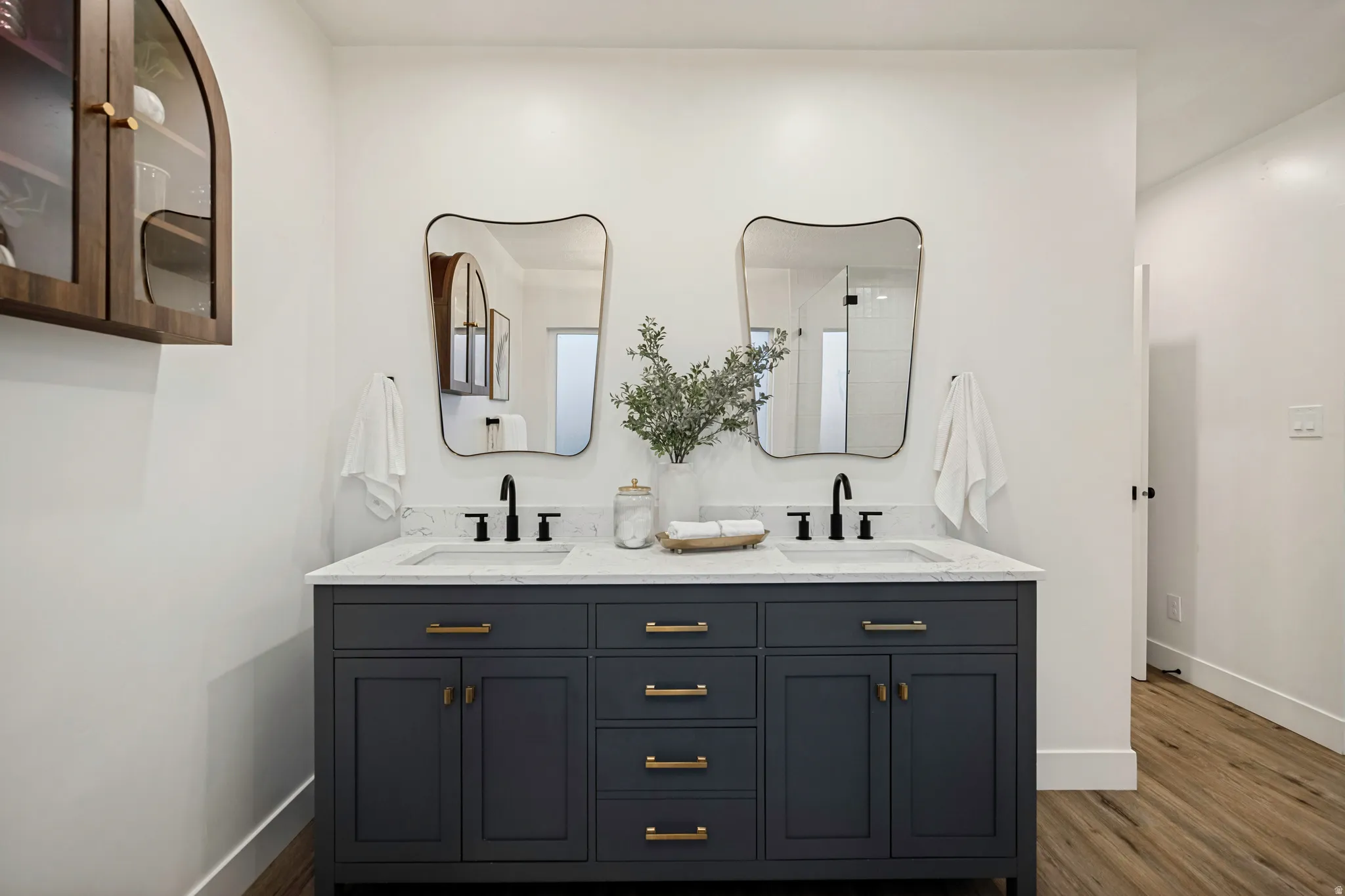 Dual vanity with a white countertop and integrated sinks, dark cabinetry with brass hardware, two irregularly shaped mirrors, and black wall-mounted faucets