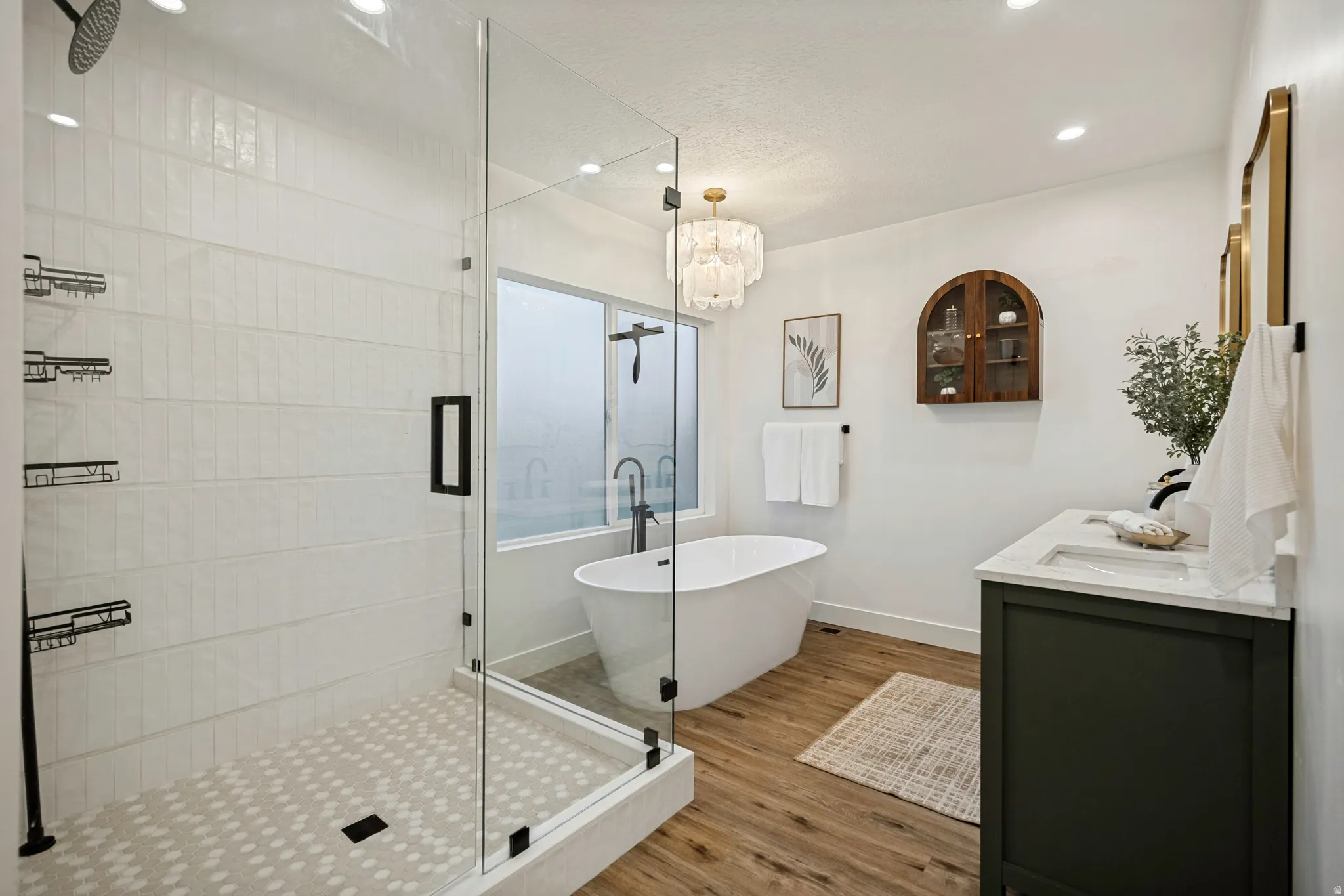 Walk-in glass shower with patterned tile floor, freestanding soaking tub, wood-finish flooring, dark green vanity with white countertop, and gold-framed mirrors