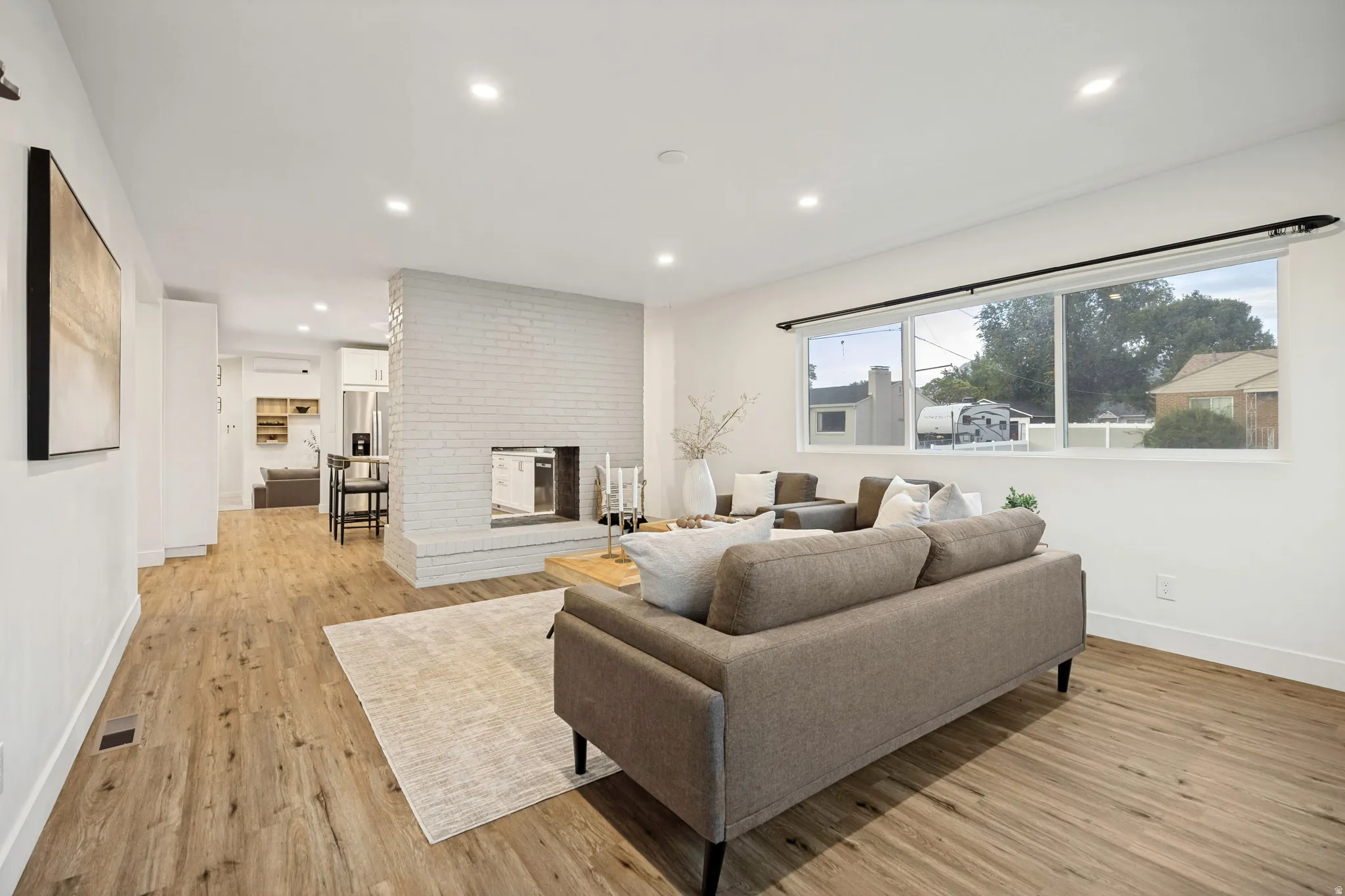 Spacious living area featuring wood-finish flooring, a white-painted brick fireplace, recessed lighting, and a large multi-pane window