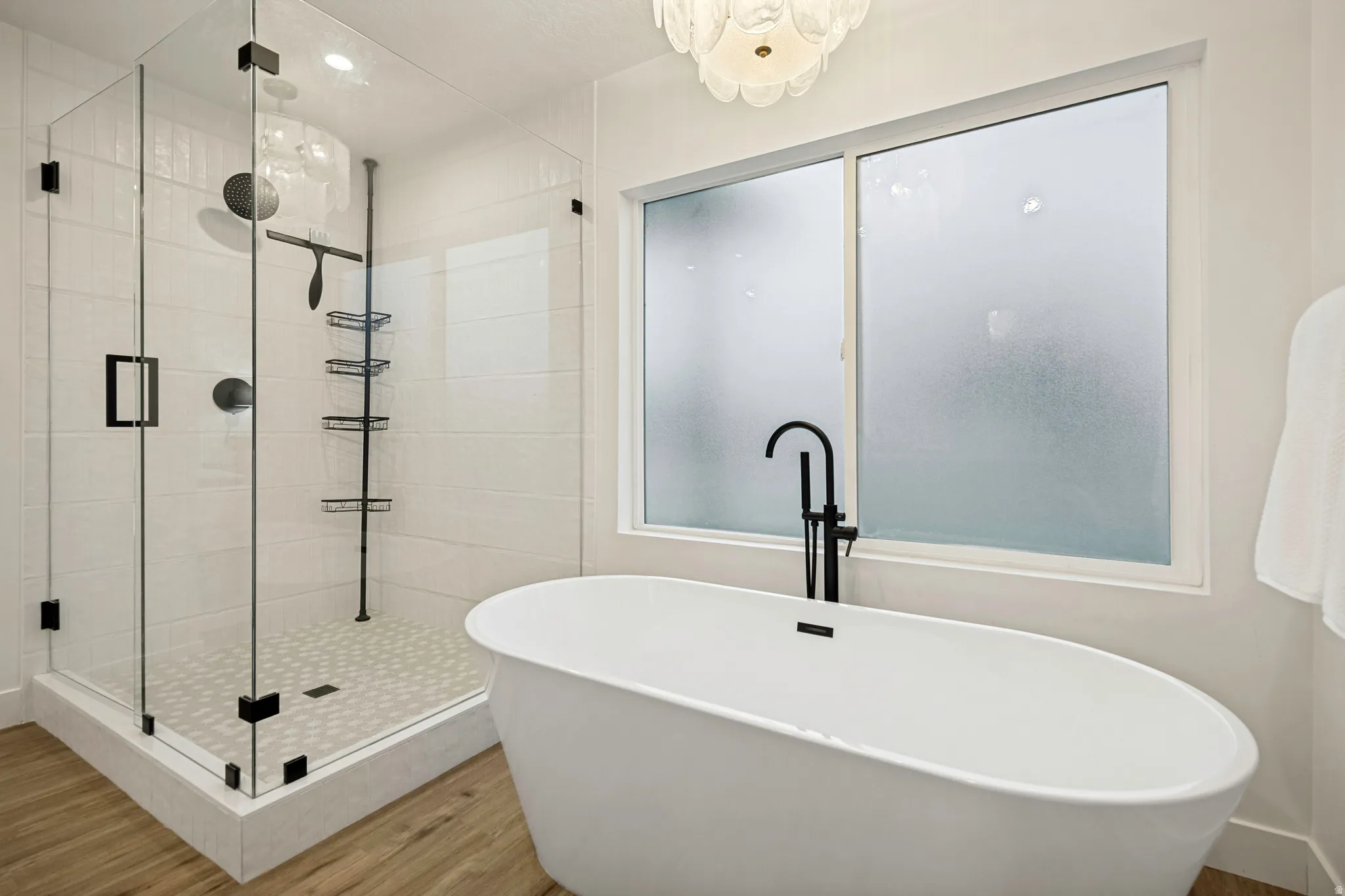 Freestanding soaking tub with a floor-mounted matte black faucet, glass-enclosed shower, rainfall showerhead, and wood-finish flooring