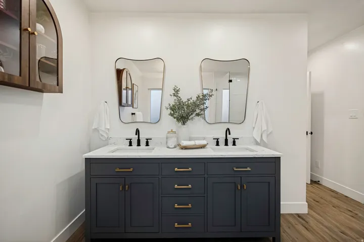 Dual vanity with a white countertop and integrated sinks, dark cabinetry with brass hardware, two irregularly shaped mirrors, and black wall-mounted faucets
