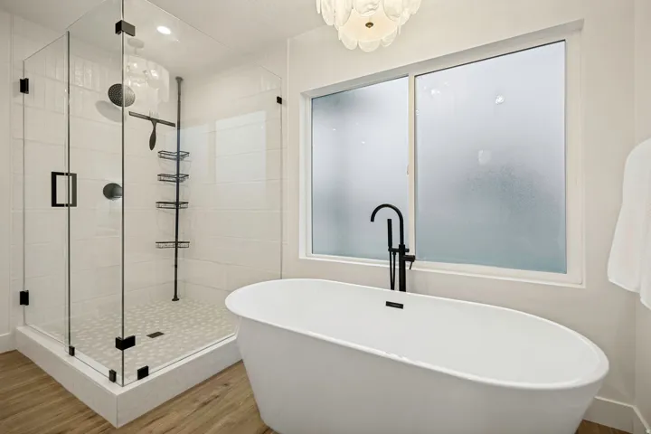 Freestanding soaking tub with a floor-mounted matte black faucet, glass-enclosed shower, rainfall showerhead, and wood-finish flooring
