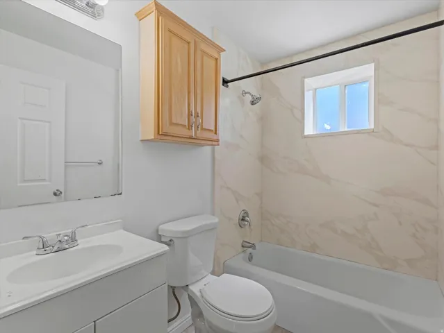 Bathroom featuring a vanity with an integrated sink, a toilet, a shower-over-tub combination with light-toned wall tile, and a window for natural light