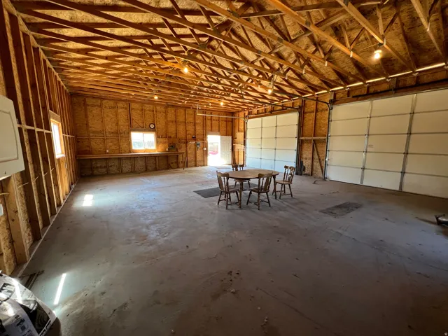Spacious garage with exposed wood beam ceiling, two large overhead doors, concrete flooring, built-in workbench, and multiple windows