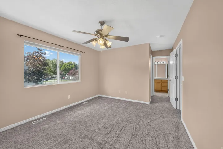 Neutral tone wall paint, gray carpeting, dual-pane window, ceiling fan with integrated lighting, and an attached bathroom featuring a wood-finish vanity with a mirror and light fixture