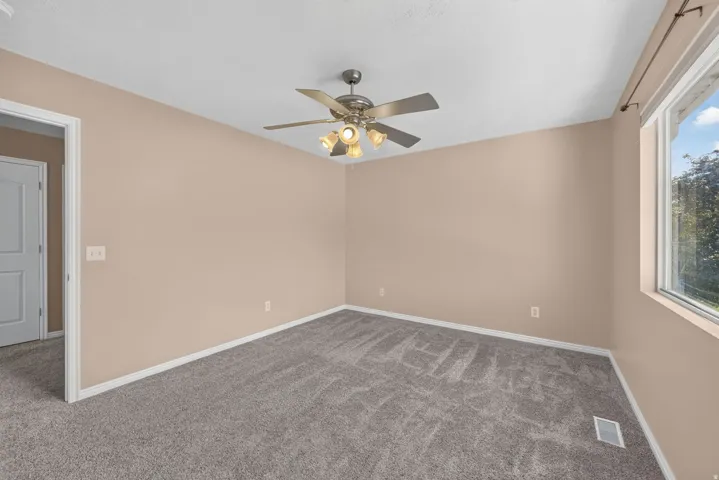 Spacious room featuring neutral carpeting, light-colored walls, a ceiling fan with integrated lighting, and a large window