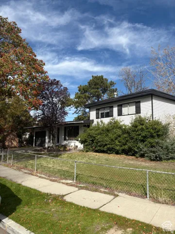 White siding exterior with dark trim and shutters, surrounded by mature trees and shrubs, featuring a front lawn with chain link fencing