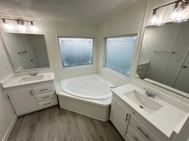 Dual vanities with integrated sinks, corner soaking tub, decorative privacy window, wood-finish flooring, and brushed nickel fixtures