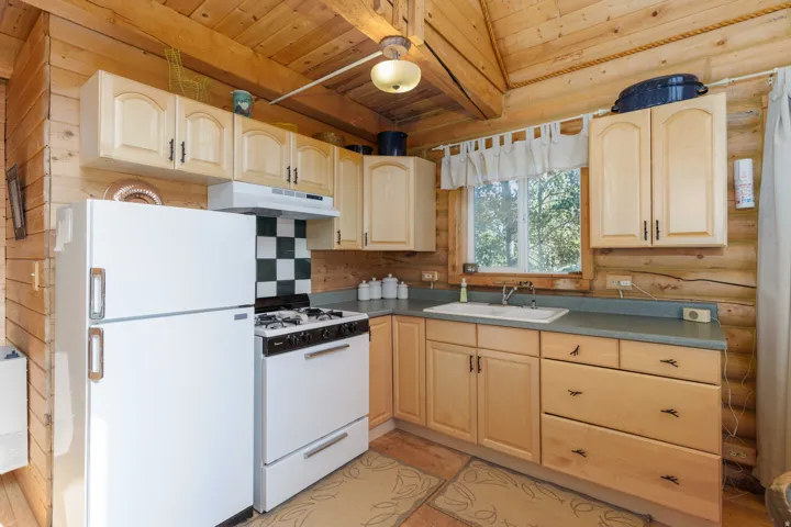 Wood-finish walls and vaulted ceiling, light-toned cabinetry with dark hardware, white appliances, checkered backsplash, and a window above the sink