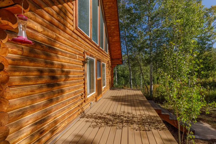 Log cabin exterior with an expansive wood-finish deck, framed by mature trees, featuring multiple windows and a glass-paneled door