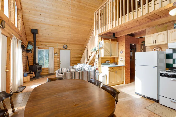 Vaulted wood-finish ceilings, an open loft area, wood-finish walls, a wood-burning stove, and a kitchen with white appliances