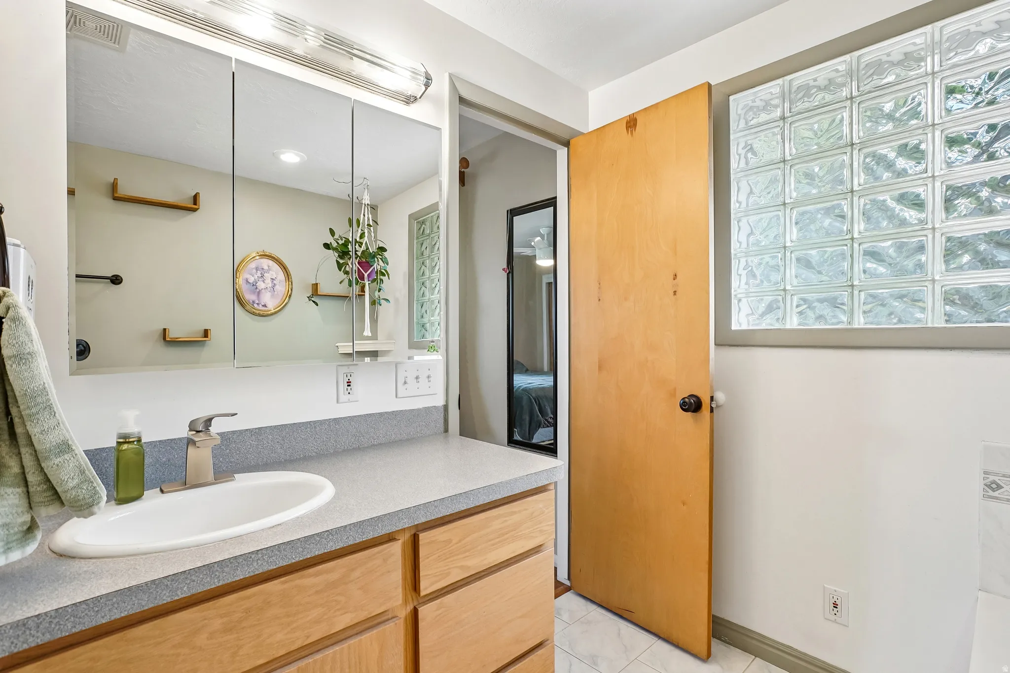 Wood-finish vanity with an integrated sink, tri-fold mirrored medicine cabinet, overhead light fixture, and glass block window