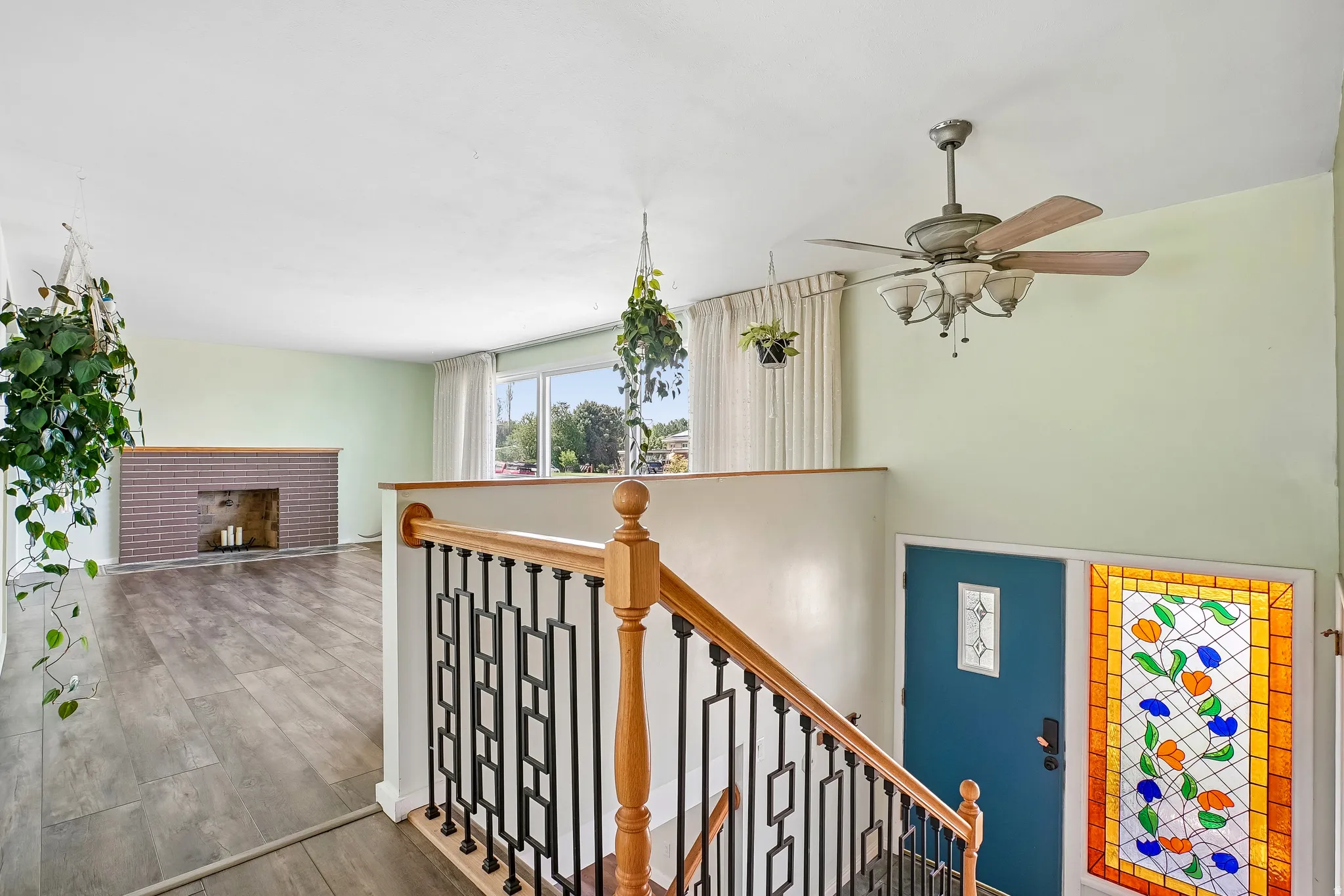 Wood-finish flooring throughout, brick fireplace with mantel, staircase with wood handrail and metal balusters, ceiling fan with light fixture, and an entry door with decorative glass