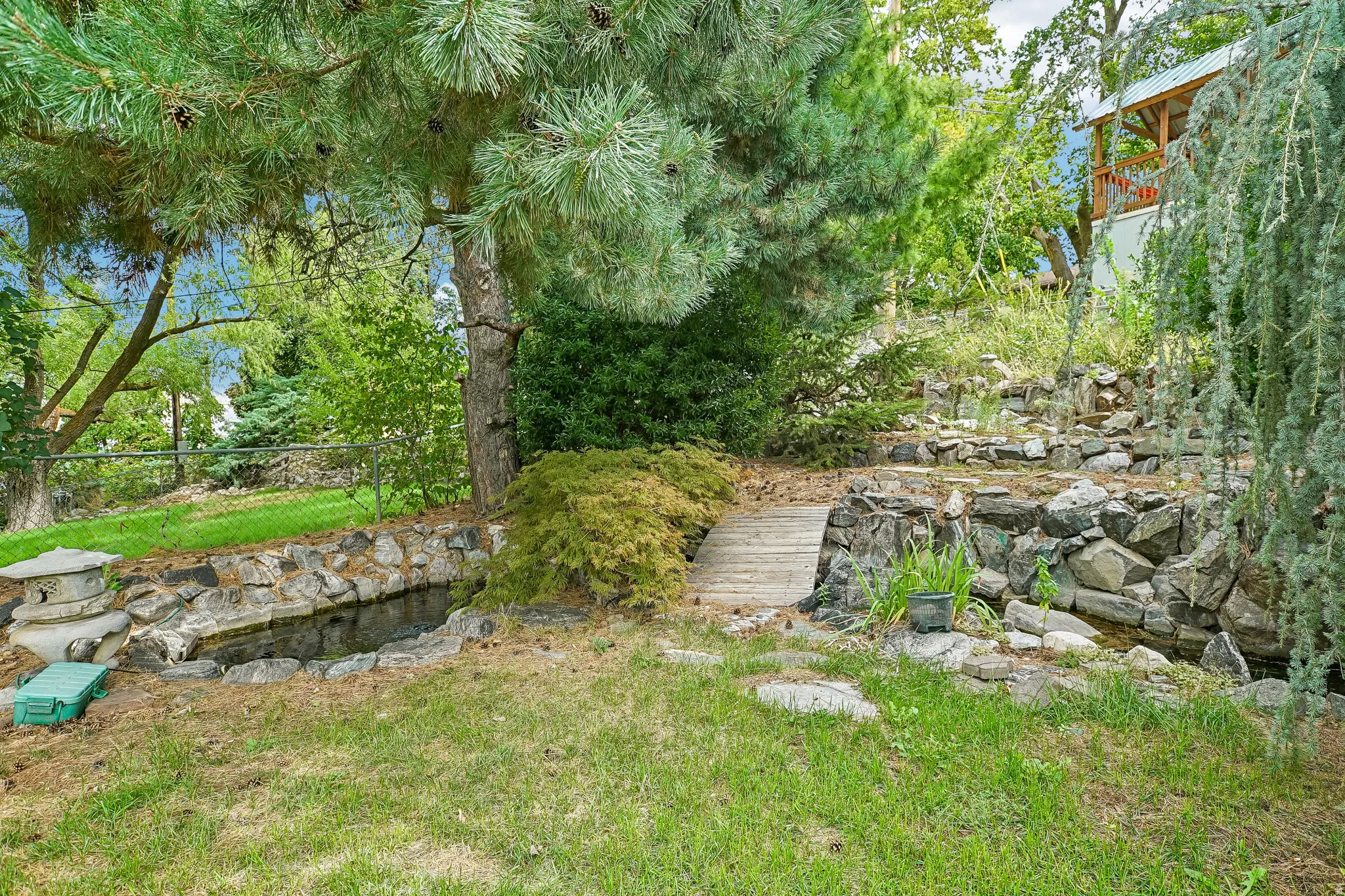 Manicured lawn with a stone-lined water feature, a small wooden bridge, and mature evergreen trees