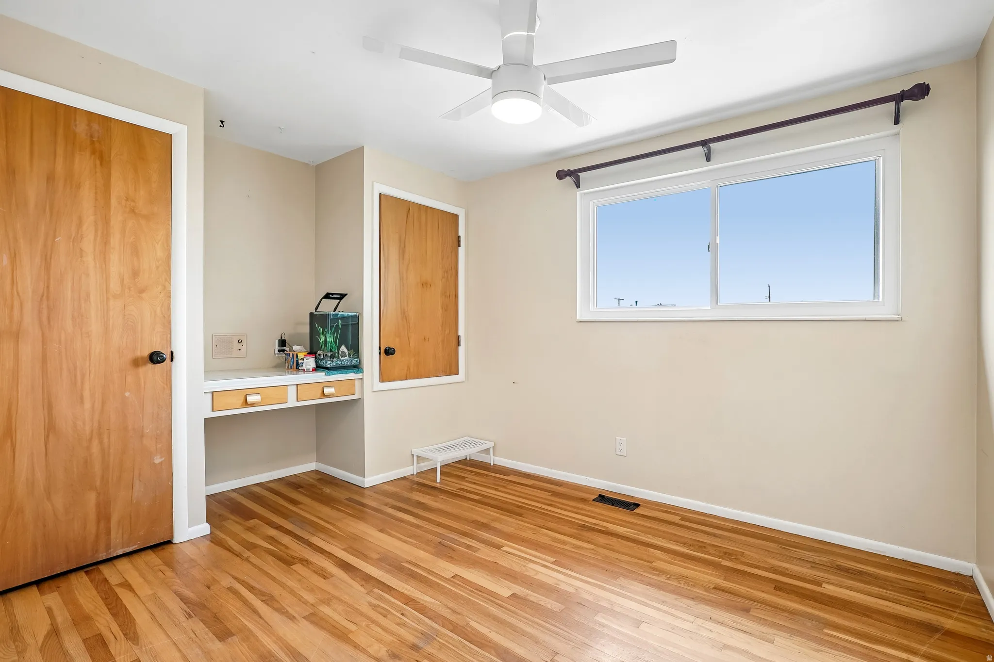 Wood-finish flooring throughout, white-framed window, built-in desk with drawers, and ceiling fan with integrated lighting