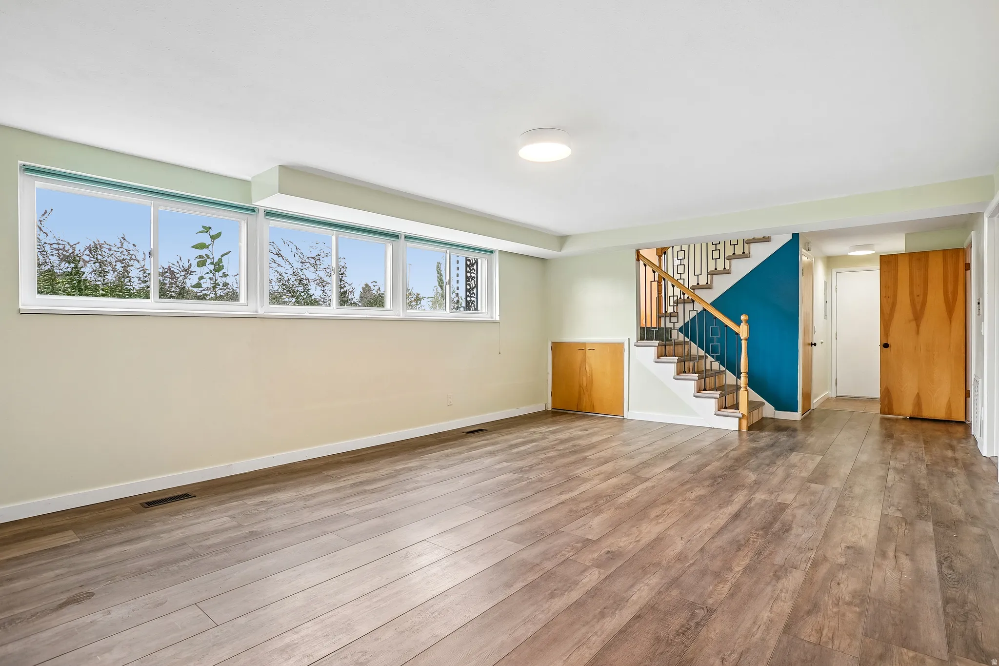 Spacious room featuring wood-finish flooring, a long window bank, a staircase with wood railings, a built-in wood-finish cabinet, and flush-mount ceiling lighting