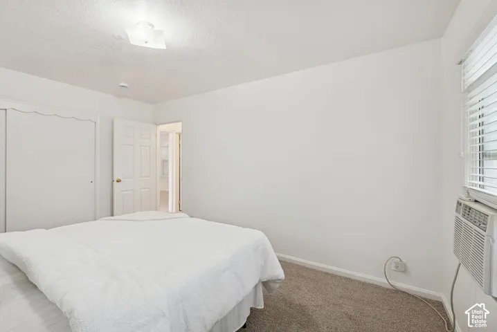 White walls with textured finish, light brown carpet, window with horizontal blinds, built-in air conditioning unit, and a built-in closet with sliding doors