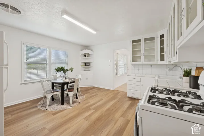 Kitchen featuring white cabinetry with glass inserts, tile backsplash, built-in corner shelving, wood-finish flooring, and a white gas range