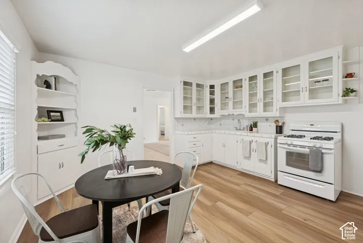 Wood-finish flooring, white cabinetry with glass inserts, white tile backsplash, built-in shelving, and a white gas range
